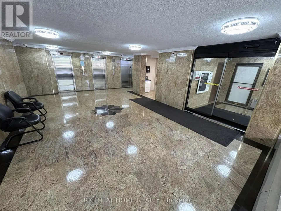 Building lobby with polished stone-look floor and walls, three metal elevator doors, black chairs and glass entrance doors.