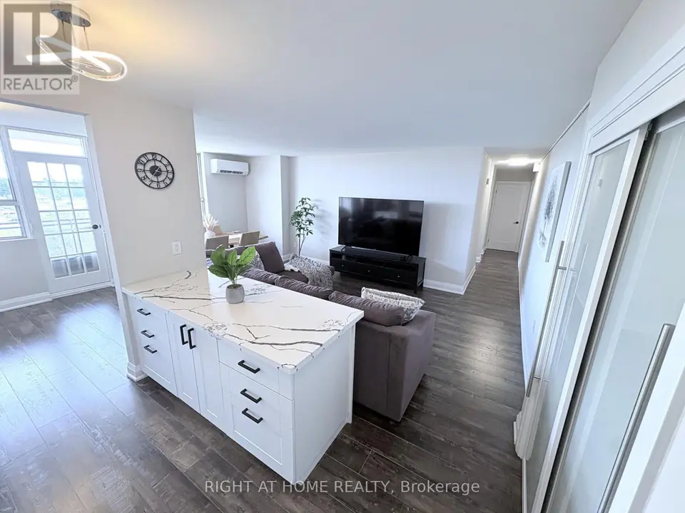 Open-concept living room with white quartz-topped kitchen peninsula, sofa, TV, dining table, wall clock and dark laminate floors.