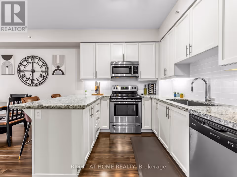 A white-cabinet kitchen seen from the dining side: a granite breakfast bar peninsula at left leads to a stainless steel range under an over-the-range microwave, then to a sink with a tall pull-down faucet and a stainless steel dishwasher at right. Lit under-cabinet lights glow over a white tile backsplash, and dark laminate flooring runs through the room.