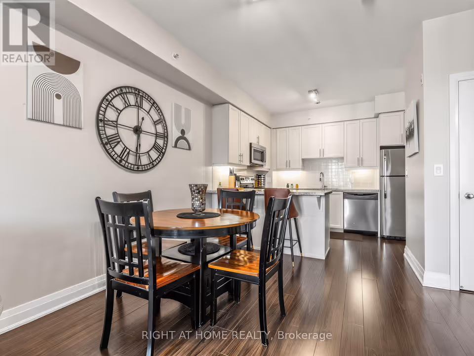 A round wooden dining table with four black chairs stands in the foreground below a large Roman-numeral wall clock and two art prints. Beyond it, white cabinets, a stainless steel microwave, a granite breakfast bar with stools, a stainless steel dishwasher and a stainless steel fridge line the kitchen. A closed white door sits at the right edge, all over dark laminate floors.