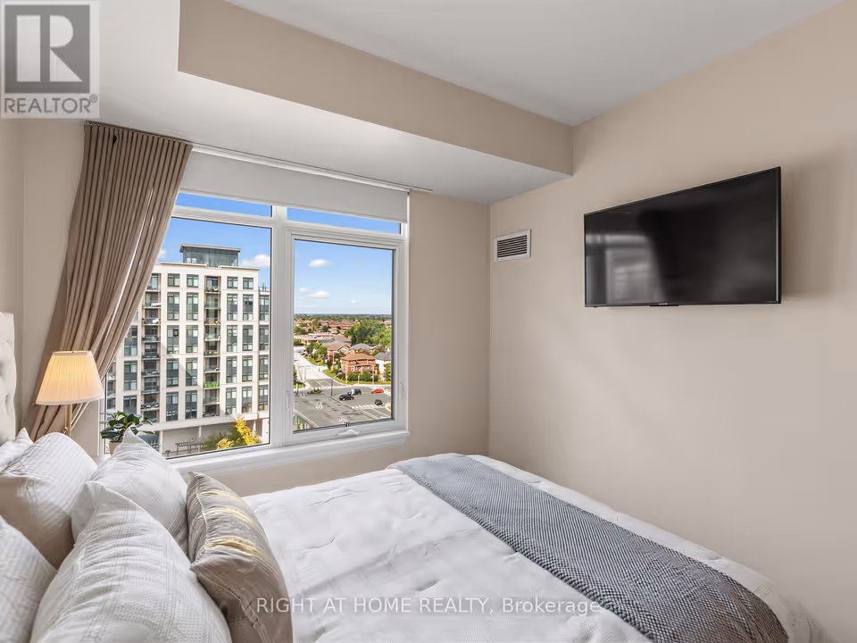 A white-framed window with a roller shade and a taupe curtain looks out at a neighbouring apartment building with balconies on the left and, beyond it, brick houses and a road intersection under a blue sky. To the right a flat-screen television is mounted on the wall beneath a stepped ceiling bulkhead, above the corner of a bed with white pillows and a grey throw.