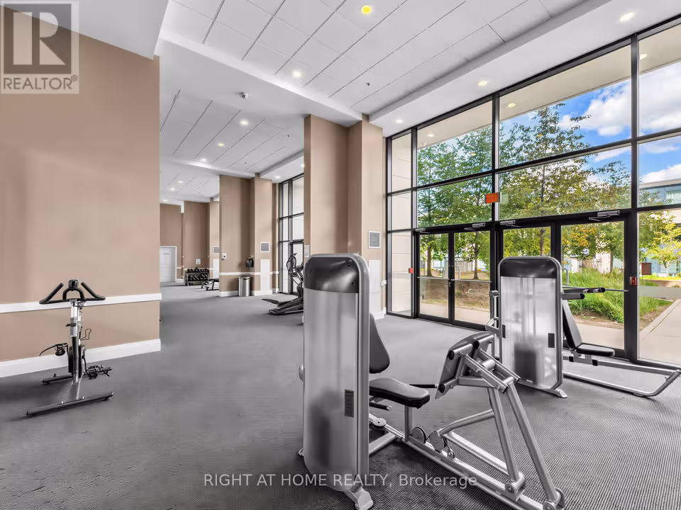 Looking along a row of beige columns in a gym with grey carpet, a stationary bike stands at left and two seated strength machines fill the foreground. At right, floor-to-ceiling windows and glass doors look out onto trees and a walkway.