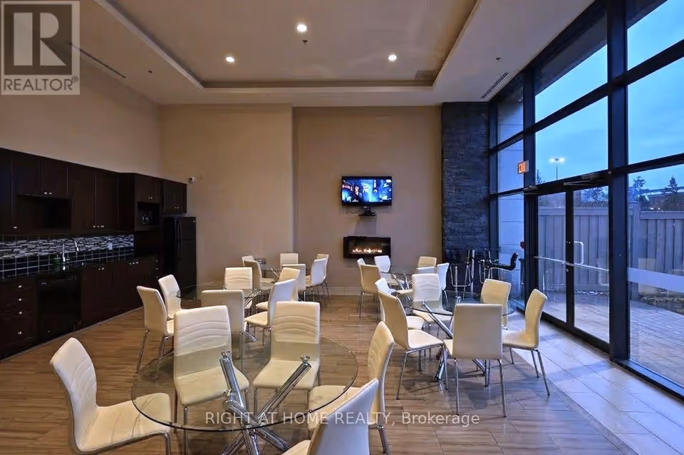 A room with a tray ceiling, wood-look floor and full-height windows at right, set with round glass-topped tables and cream chairs. Dark cabinetry with a sink and a black fridge runs along the left wall, and a wall-mounted TV sits above a linear fireplace beside a stacked-stone wall section.